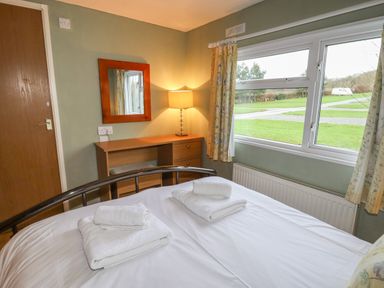 A bedroom with a bed, desk with a lamp, mirror on the wall, window with curtains, and towels on the bed at Wayside Lodge in Aston On Clun