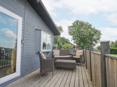 An outdoor wooden deck with wicker chairs sofa and table attached to a house at Wayside Lodge in Aston On Clun