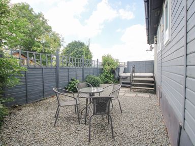 An outdoor gravel area with four chairs and a round glass table next to a building with stairs leading to a hot tub at Wayside Lodge in Aston On Clun