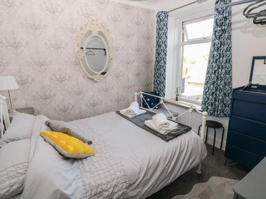 A bedroom with a bed with folded towels on it a blue chest of drawers window with patterned curtains and an ornate oval mirror on the wall at 5 Pontardulais Road in Llangennech