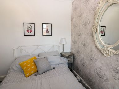 A bedroom with a bed gray bedding colorful pillows two framed pictures a nightstand with a lamp and a round decorative mirror on the wall at 5 Pontardulais Road in Llangennech