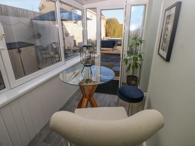 A small glass table with a decorative candle holder between a cream chair and a blue stool in a sunroom with windows and a door leading outside at 5 Pontardulais Road in Llangennech