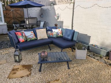 An outdoor seating area with a blue corner sofa with cushions a small table with a lantern and a patio table with chairs and an umbrella at 5 Pontardulais Road in Llangennech