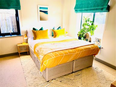 A bedroom with a bed covered with yellow bedding and green pillows next to two windows with green blinds at Avalennek in St Newlyn East