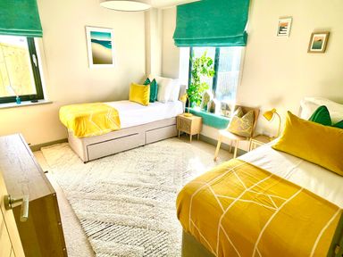 A bedroom with two single beds yellow blankets green cushions bedside tables and window seats at Avalennek in St Newlyn East