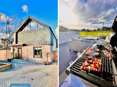 An exterior view of a modern house with wood and shingle siding and a paved driveway and a barbecue grill on a deck with cooking meat and outdoor seating with a view at Avalennek in St Newlyn East