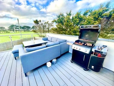 An outdoor patio with a blue sectional sofa around a table and a gas grill with food on the grill at Avalennek in St Newlyn East
