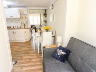 A small living and dining area with a gray sofa and decorative pillow next to a glass dining table with white chairs and a kitchen in the background at Y Bwthyn Bach in Tregarth