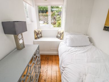 A small bedroom with a single bed a gray dresser with a lamp and a window seat with animal print pillows at Y Bwthyn Bach in Tregarth