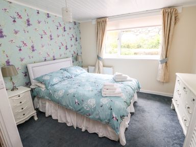 A bedroom with a bed with blue floral bedding two bedside tables lamps a window with curtains and a white dresser at Y Bwthyn Bach in Tregarth