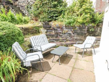An outdoor patio area with a glass table and three striped cushioned chairs surrounded by stone walls and greenery at Y Bwthyn Bach in Tregarth