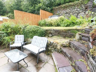 An outdoor patio area with striped cushioned chairs and a glass table next to stone steps and plants at Y Bwthyn Bach in Tregarth