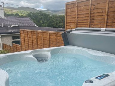 An outdoor hot tub with bubbling water next to wooden fences and a house roof with hills and trees in the background at Y Bwthyn Bach in Tregarth