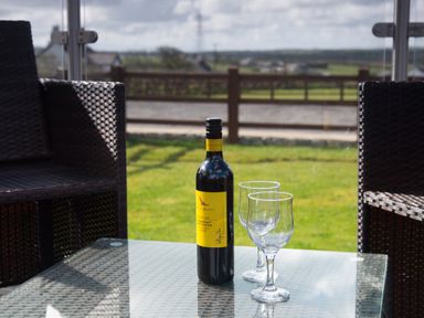 A glass table with a bottle of wine and two empty wine glasses between two wicker chairs on a patio overlooking a lawn at The Lodge in Rhosybol