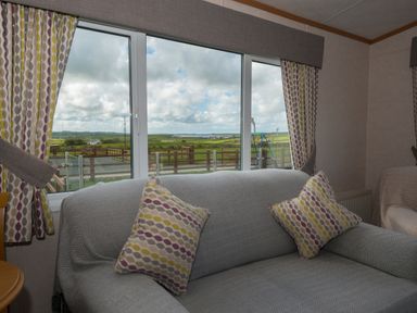 A living room with a sofa and patterned cushions in front of large windows with a view of fields at The Lodge in Rhosybol
