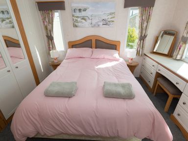 A bedroom with a double bed covered with pink bedding and two folded towels two bedside tables with lamps a dressing table with a mirror and stool and floral curtains at The Lodge in Rhosybol