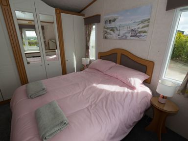A bedroom with a double bed pink bedding two folded towels bedside table with lamp wardrobe with mirrored doors and a window at The Lodge in Rhosybol