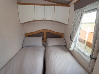 A bedroom with two single beds with grey bedding under white wall cabinets and a window with curtains at The Lodge in Rhosybol