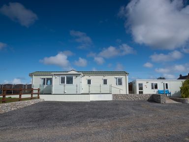 A gravel parking area in front of two light-colored modular buildings with glass railings and a blue bench at The Lodge in Rhosybol