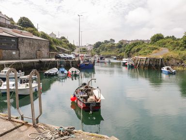 A harbor with multiple boats docked along stone piers and cars parked near a stone building with greenery on the hills at The Lodge in Rhosybol