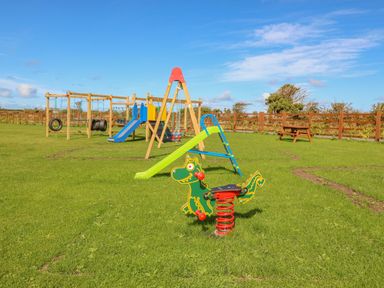 A playground with slides swings and a spring rider on grass with a picnic table and fence in the background at The Lodge in Rhosybol