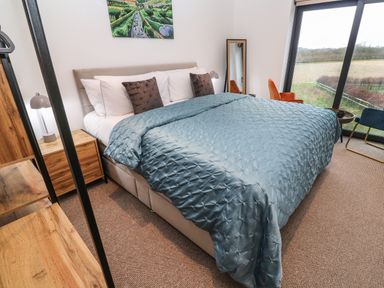 A bedroom with a large bed covered in a blue quilt two brown pillows a wooden nightstand with a lamp a full-length mirror two chairs and a window showing a field at Tredarn in Connor Downs