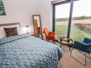 A bedroom with a bed blue cover and two chairs around a round wooden table near a large window at Tredarn in Connor Downs