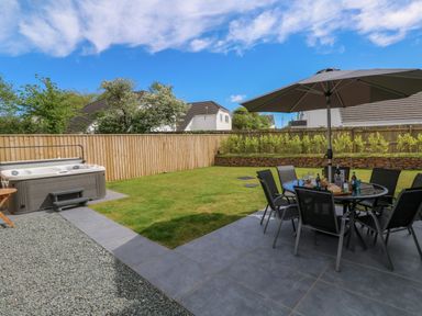 A backyard with a hot tub a dining table with chairs and an umbrella on a patio surrounded by a lawn a wooden fence and houses at Tredarn in Connor Downs