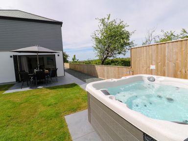 An outdoor patio area with a hot tub a dining table with chairs and an umbrella a wooden fence and a grassy lawn at Tredarn in Connor Downs