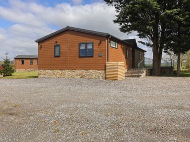 A brown wooden building with stone base on a gravel lot with trees and another building in the background at Ruperts Retreat in Nateby near Garstang