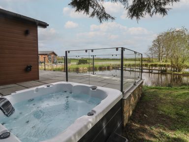 An outdoor patio with a hot tub next to a wooden building and a pond with trees at Ruperts Retreat in Nateby near Garstang