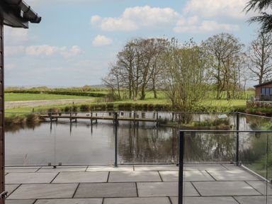 A view of a pond with a wooden bridge and trees behind a glass railing on a stone patio at Ruperts Retreat Nateby near Garstang