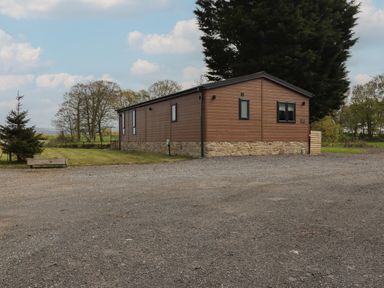 A brown single-story building with stone base surrounded by gravel and grass with trees in the background at Ruperts Retreat in Nateby near Garstang