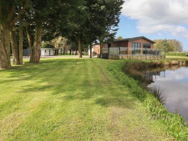 A grassy area with trees next to a pond and a house with a glass railing at Ruperts Retreat in Nateby near Garstang