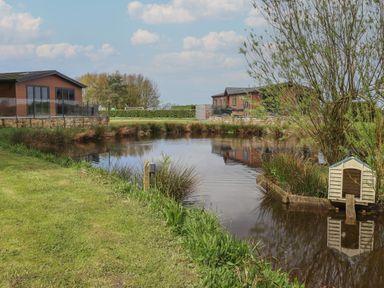 A pond with a small wooden house on the edge and two wooden cabins in the background at Ruperts Retreat in Nateby near Garstang