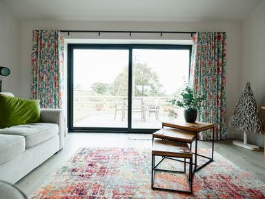 A living room with a gray sofa and green cushion floral curtains glass sliding doors with outdoor furniture and nested wooden tables with a plant on a colorful rug at Ingleborough Lodge in Rathmell near Settle