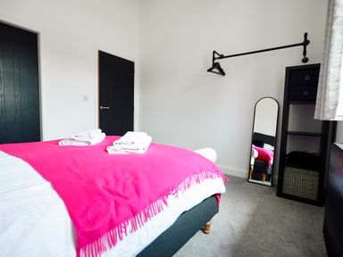 A bedroom with a bed covered by a pink blanket with towels on it a tall mirror a black shelving unit with drawers two black doors and a hanging rod with hangers at Ingleborough Lodge in Rathmell near Settle