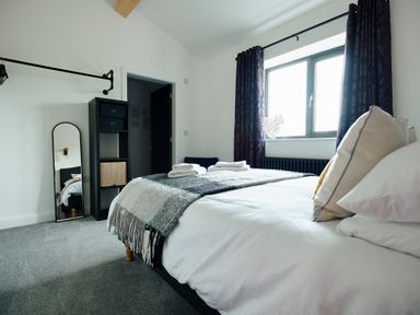 A bedroom with a bed with pillows and folded towels a window with dark curtains a standing mirror and a shelving unit at Ingleborough Lodge in Rathmell near Settle