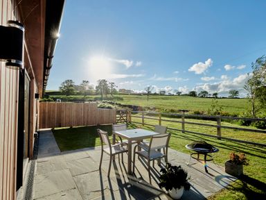 A patio with a table and four chairs a fire pit and plants overlooking a fenced grassy field at Ingleborough Lodge in Rathmell near Settle