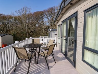 An outdoor deck with a glass table and four chairs attached to a beige mobile home with white railing and trees in the background at Crosswinds 68 in Bembridge