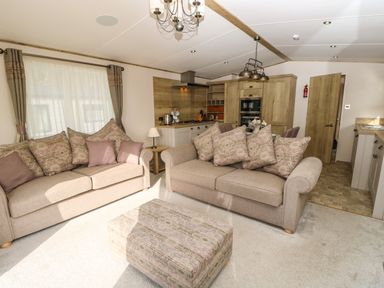 A living area with two beige sofas and a patterned ottoman near an open kitchen with wooden cabinets at Crosswinds 68 in Bembridge