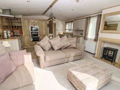 A living room with sofas and cushions a patterned ottoman fireplace and an open kitchen with wooden cabinets at Crosswinds 68 in Bembridge