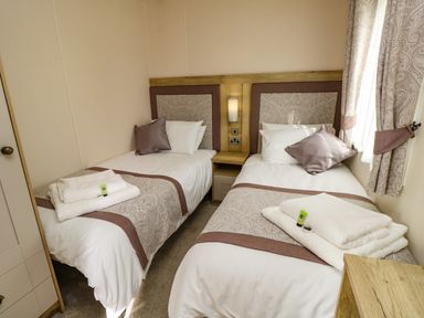A bedroom with two single beds with patterned bed runners and pillows a nightstand between the beds and curtains on the window at Crosswinds 68 in Bembridge