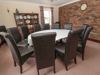 A dining room with a large oval table and dark brown chairs with a wooden cabinet displaying plates and a brick wall with a clock at Barnacre in Longhirst near Morpeth