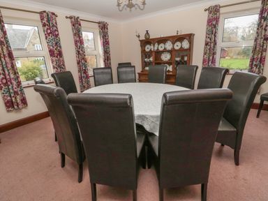 A dining room with a round table covered with a cloth and surrounded by black chairs and windows with floral curtains at Barnacre Longhirst near Morpeth