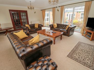 A living room with three brown leather sofas a wooden coffee table a TV on a stand and large windows with curtains at Barnacre in Longhirst near Morpeth