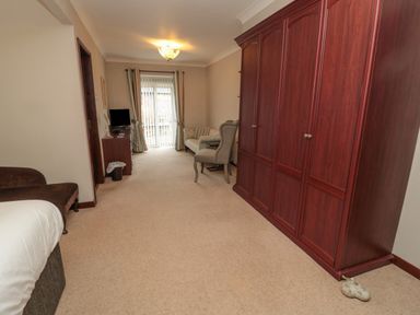 A room with a large wooden wardrobe a TV on a desk a sofa an armchair and a glass door with curtains at Barnacre in Longhirst near Morpeth