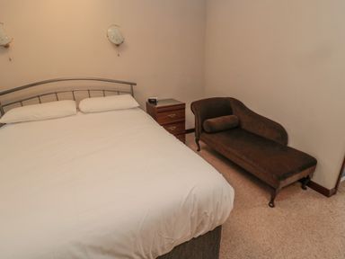 A bedroom with a bed white bedding a wooden bedside table with an alarm clock and a brown chaise lounge at Barnacre Longhirst near Morpeth