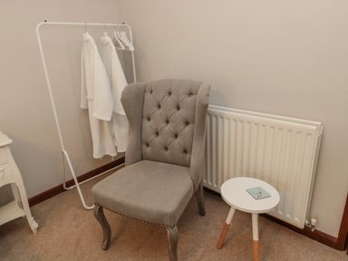 A chair next to a radiator and small round table with a book and a clothing rack with robes in a room at Barnacre in Longhirst near Morpeth