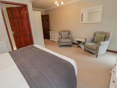 A bedroom with a bed two armchairs a white side table a chest of drawers and wooden doors at Barnacre in Longhirst near Morpeth